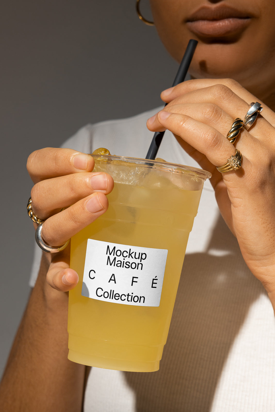 PSD mockup of a person holding a clear cup with a black straw, wearing multiple rings and a white shirt, labeled "Mockup Maison Café Collection."