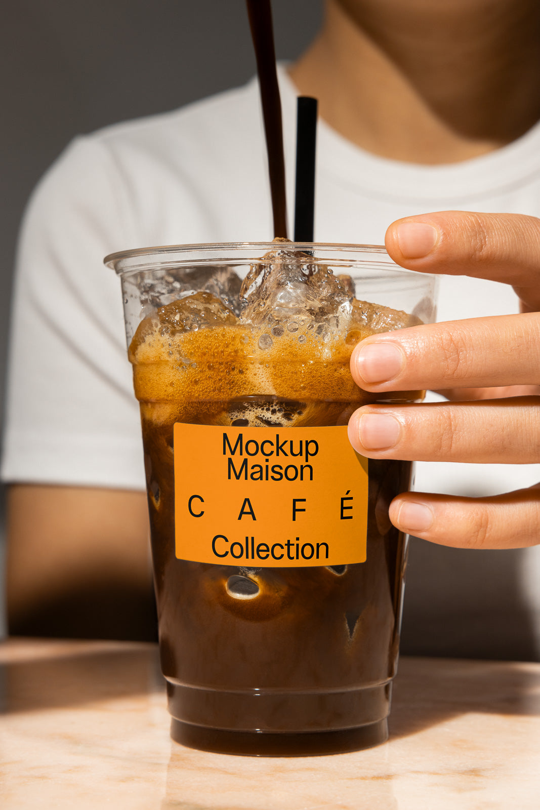PSD mockup of a person holding a clear plastic cup filled with iced coffee, featuring a label that reads "Mockup Maison Café Collection."