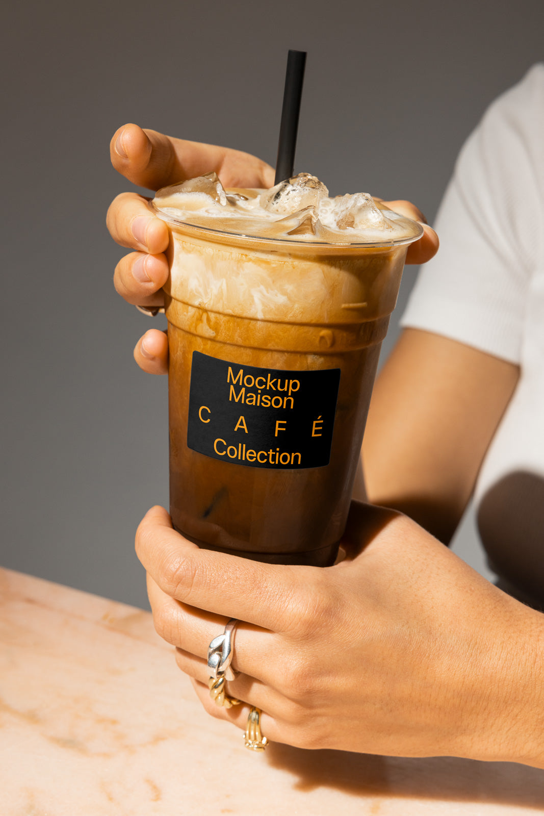 PSD mockup of a hand holding an iced coffee in a plastic cup with a black straw, labeled "Mockup Maison Café Collection" on a marble surface.