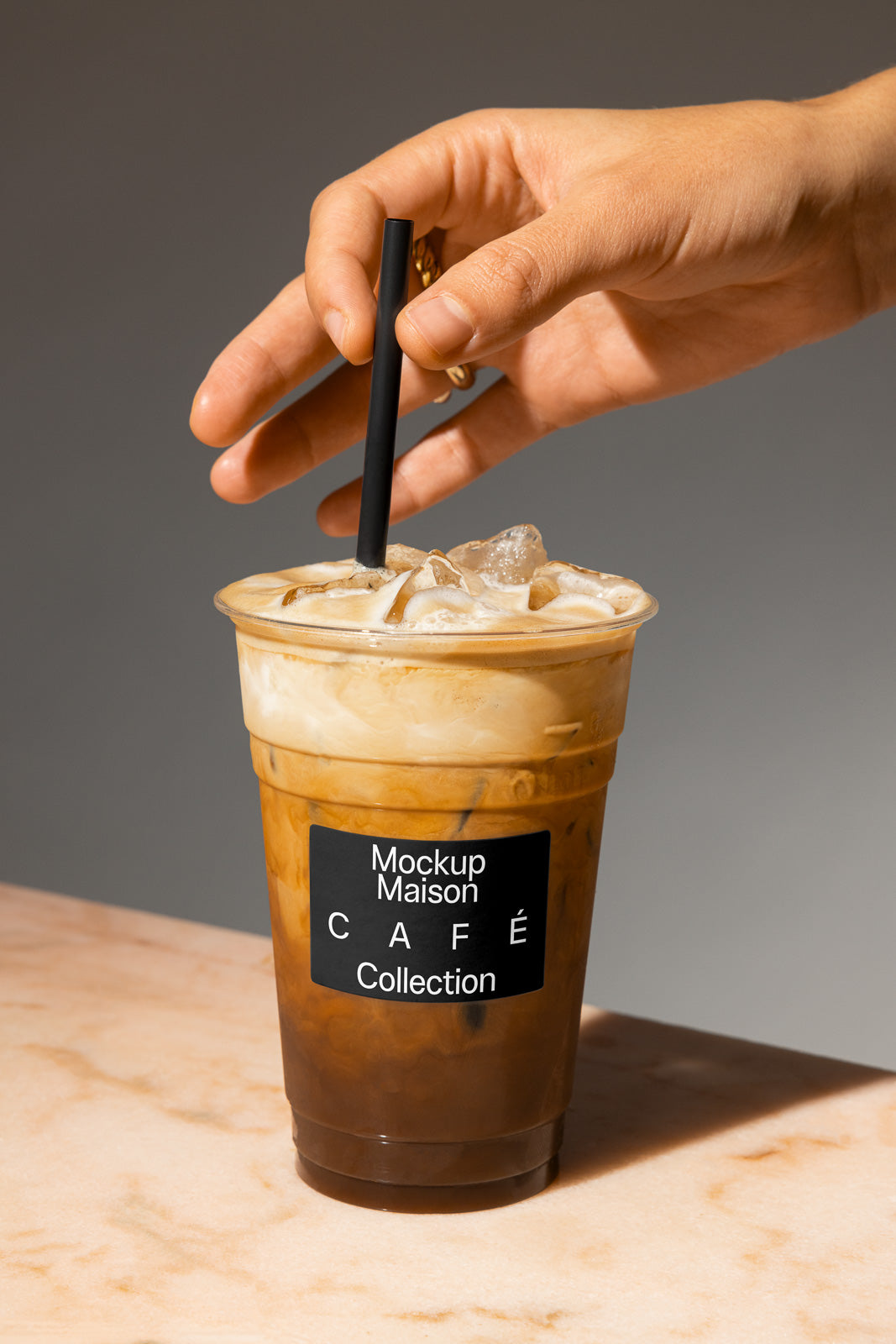 PSD mockup of a hand holding a plastic cup filled with iced coffee and a black straw, sitting on a marble surface, labeled "Mockup Maison Café Collection."