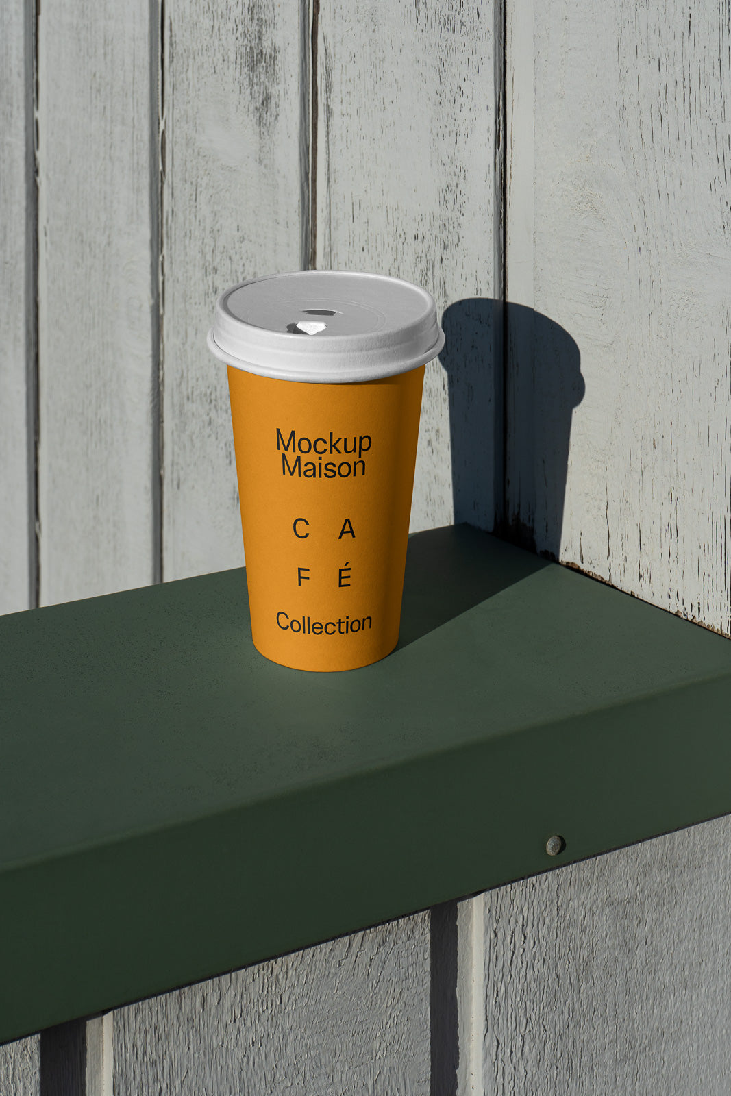 PSD mockup of a yellow paper coffee cup with white lid on a green shelf, featuring "Mockup Maison Café Collection" in black text. Cup casts a shadow.