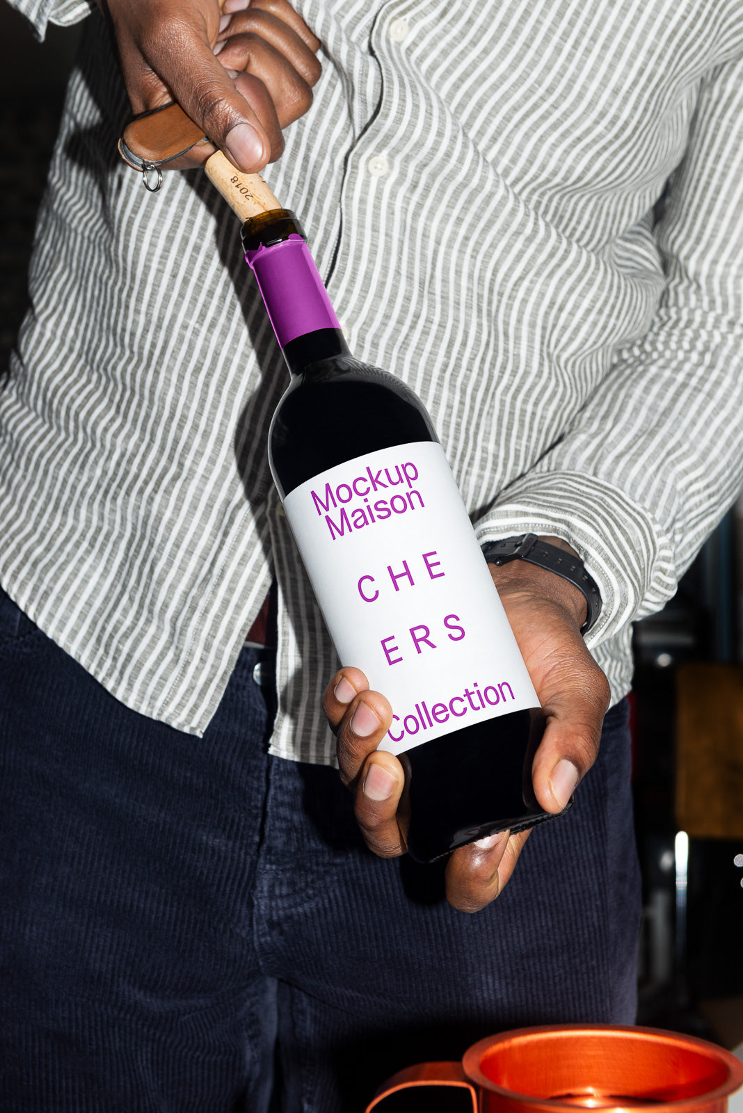 PSD mockup of a hand holding a wine bottle with a label reading "Mockup Maison Cheers Collection," next to an orange mug.