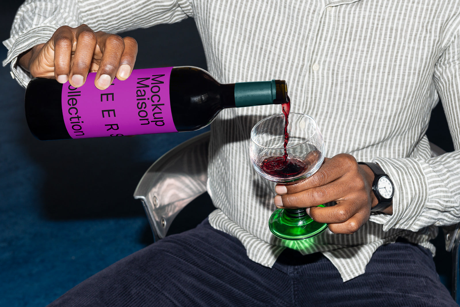 PSD mockup of a person pouring red wine from a bottle with a purple label into a stemmed glass, wearing a striped shirt and wristwatch.