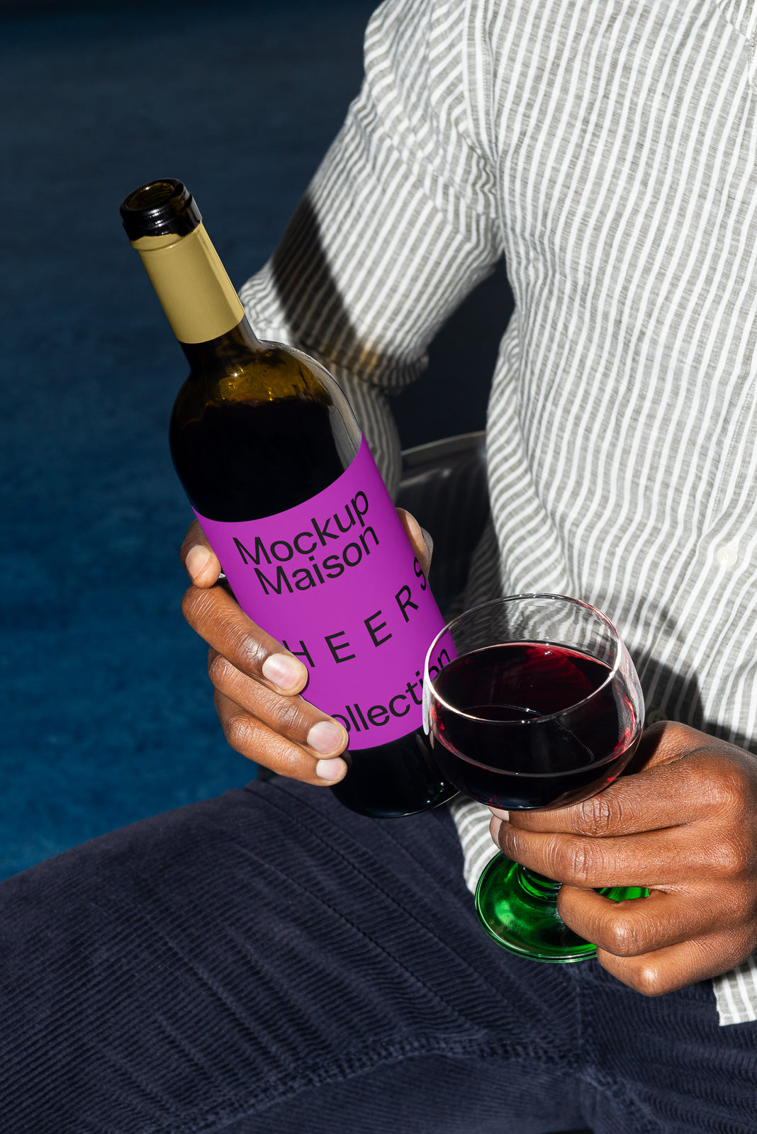 PSD mockup of a person holding a wine bottle with a purple label and pouring wine into a glass, seated on a blue surface.
