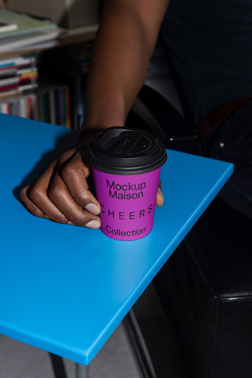 PSD mockup of a person holding a purple takeaway coffee cup labeled "Mockup Maison Cheers Collection," resting on a bright blue table surface.