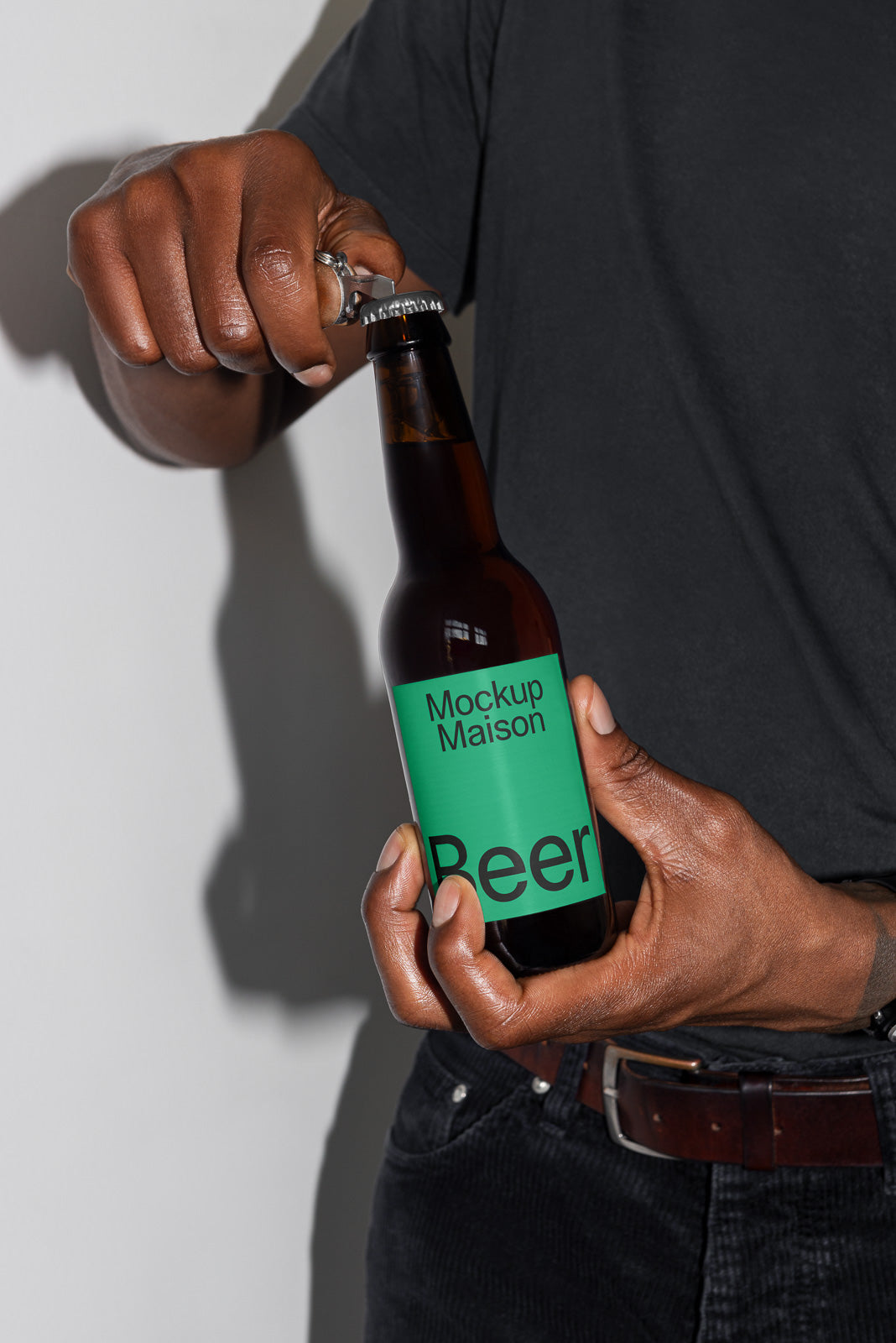 PSD mockup of a person opening a brown beer bottle labeled "Mockup Maison Beer" with a green label, wearing a black shirt and using a silver bottle opener.