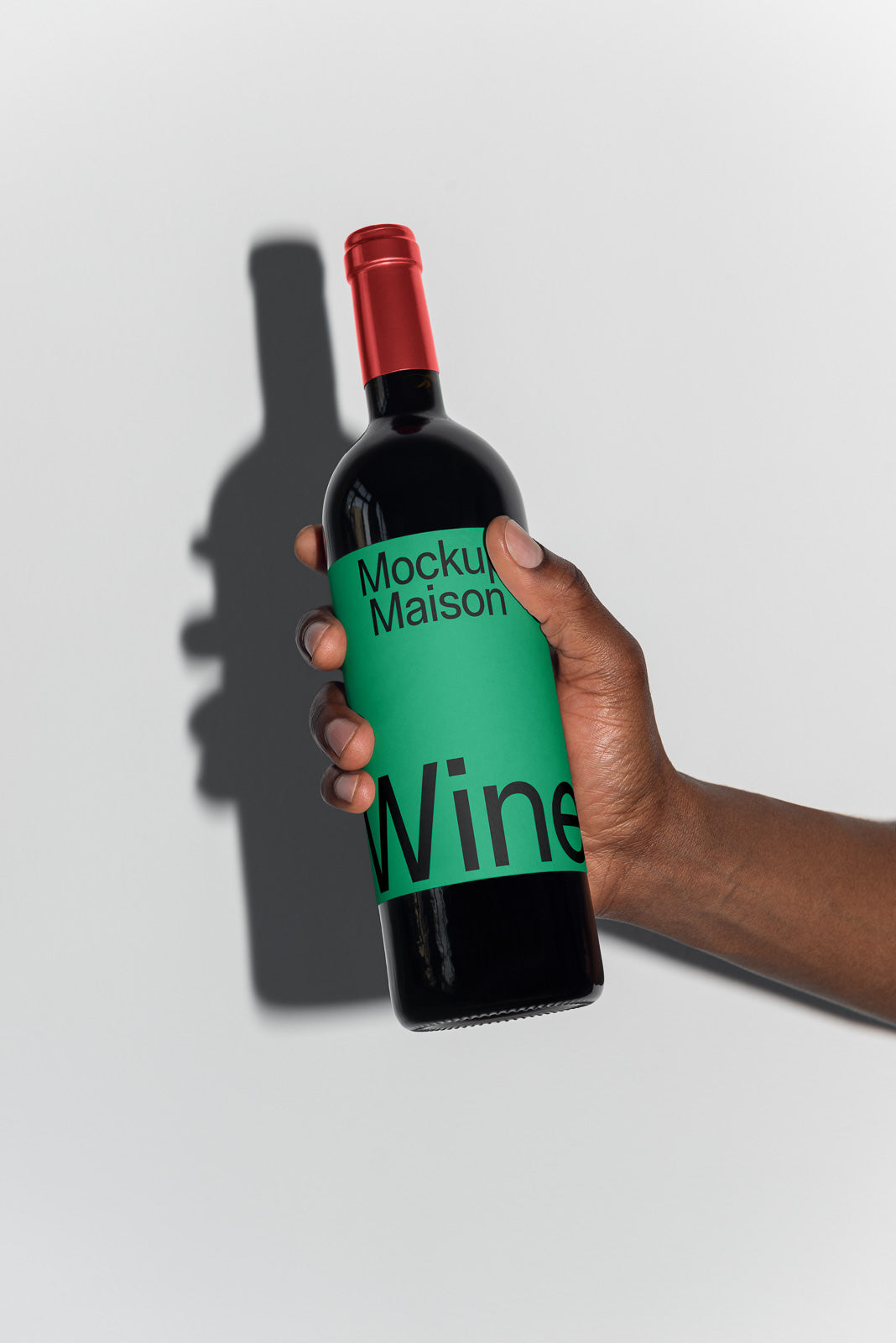 PSD mockup of a hand holding a wine bottle with a green label and red cap against a light background, casting a shadow on a surface.