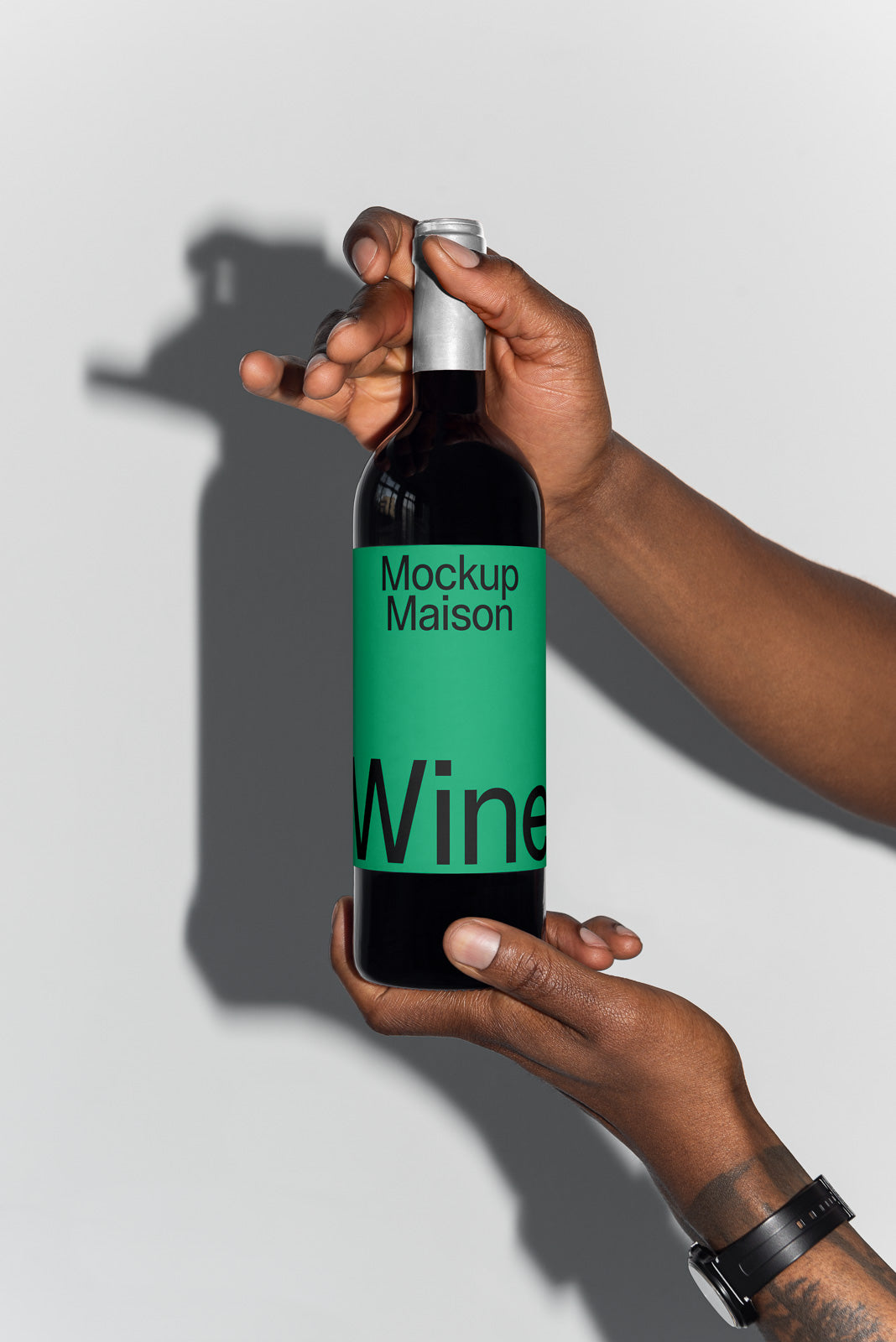 PSD mockup of a wine bottle held by hands, featuring a green label with "Mockup Maison" and black text on a silver cap against a gray wall.