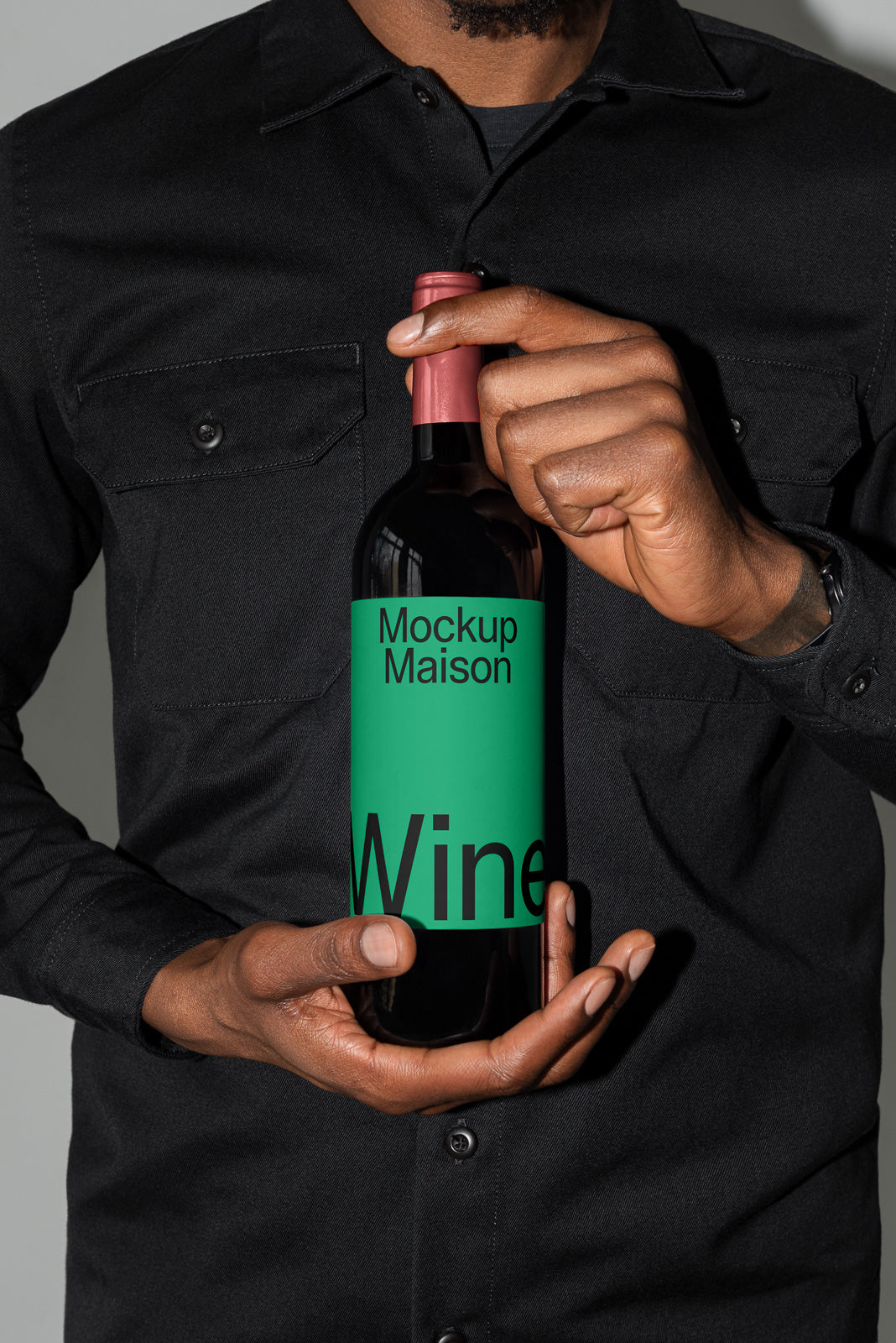 PSD mockup of a person holding a wine bottle labeled "Mockup Maison Wine" with a green label and red cap, wearing a black shirt.