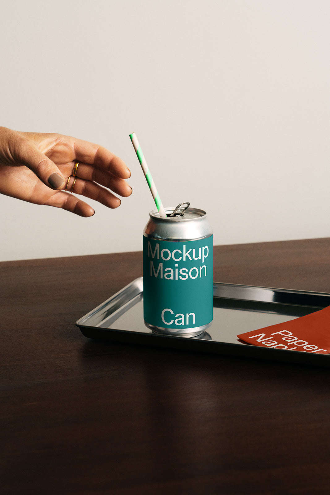 PSD mockup of a can with a teal and white design, placed on a metal tray with a red napkin. A hand reaches towards it with a striped straw inside.