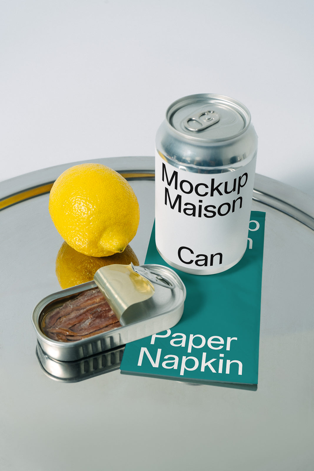 PSD mockup of a silver can labeled "Mockup Maison Can" on a reflective surface with a lemon, an open tin of anchovies, and a green paper napkin.