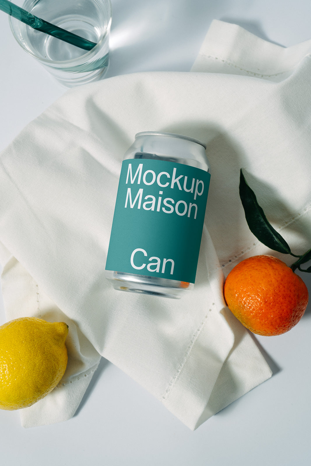 PSD mockup of a beverage can with a teal label lying on a white cloth, surrounded by a lemon, orange, and a glass with a straw.