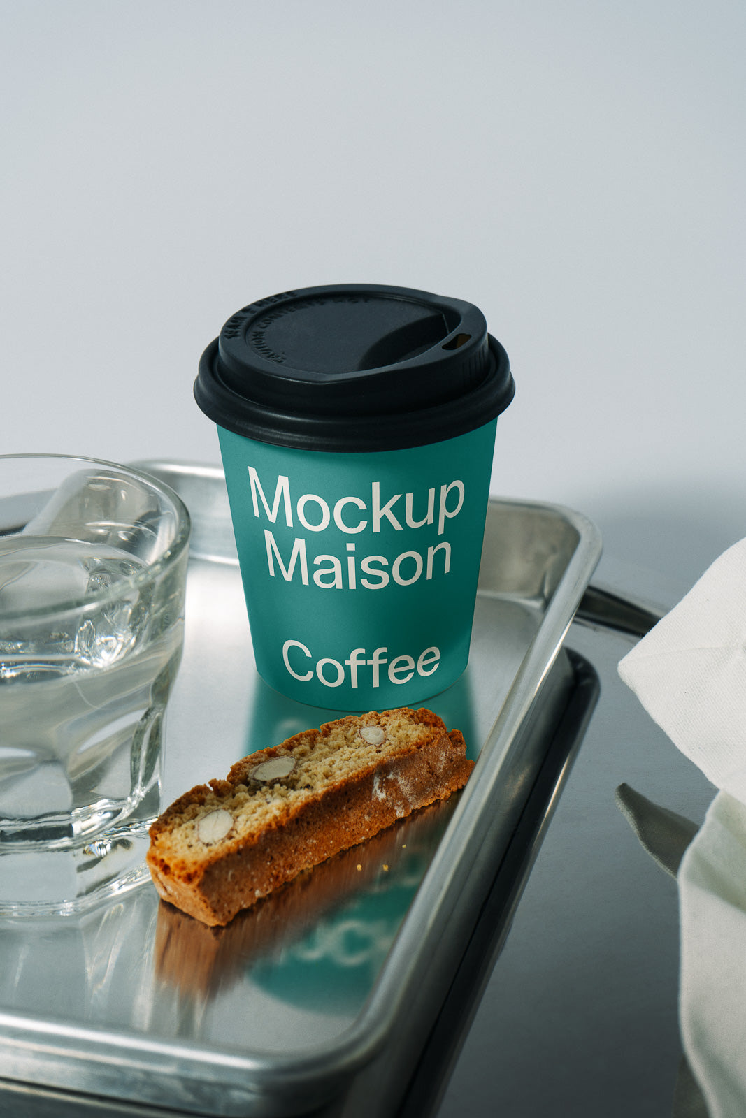 PSD mockup of a green coffee cup labeled "Mockup Maison Coffee" with a black lid on a metallic tray beside a glass of water and biscotti.