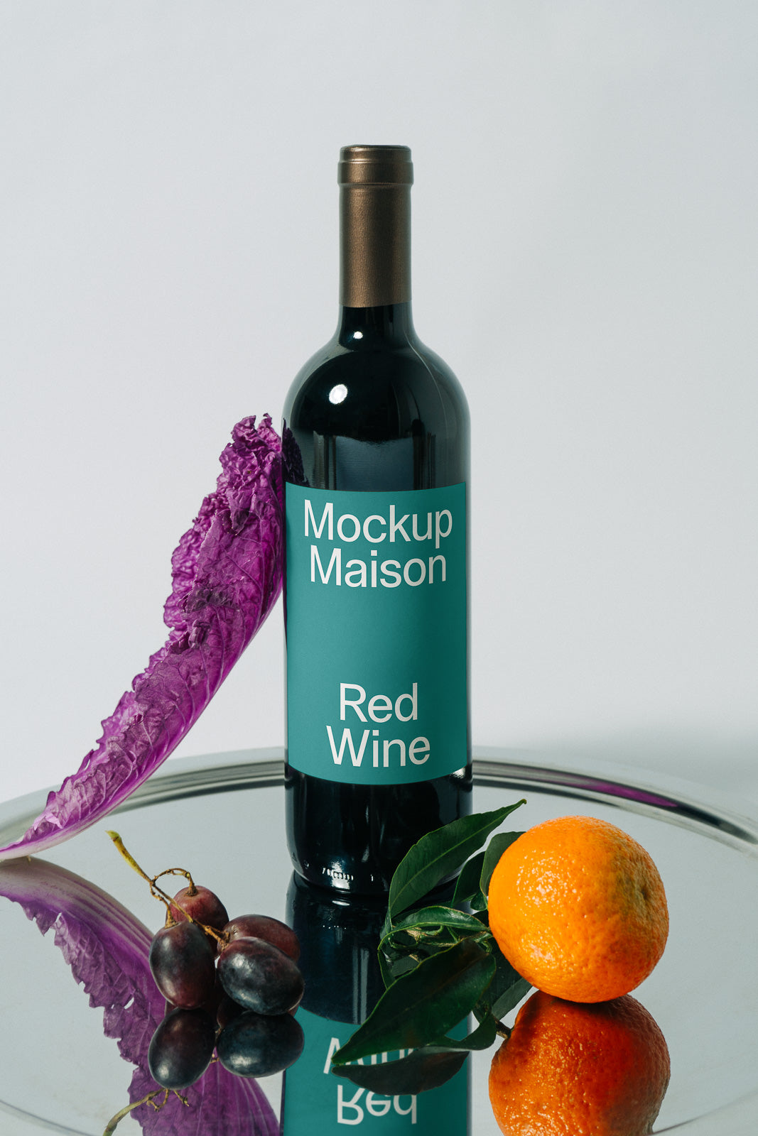 PSD mockup of a wine bottle labeled "Mockup Maison Red Wine" on a reflective surface, accompanied by a purple cabbage leaf, grapes, and an orange.