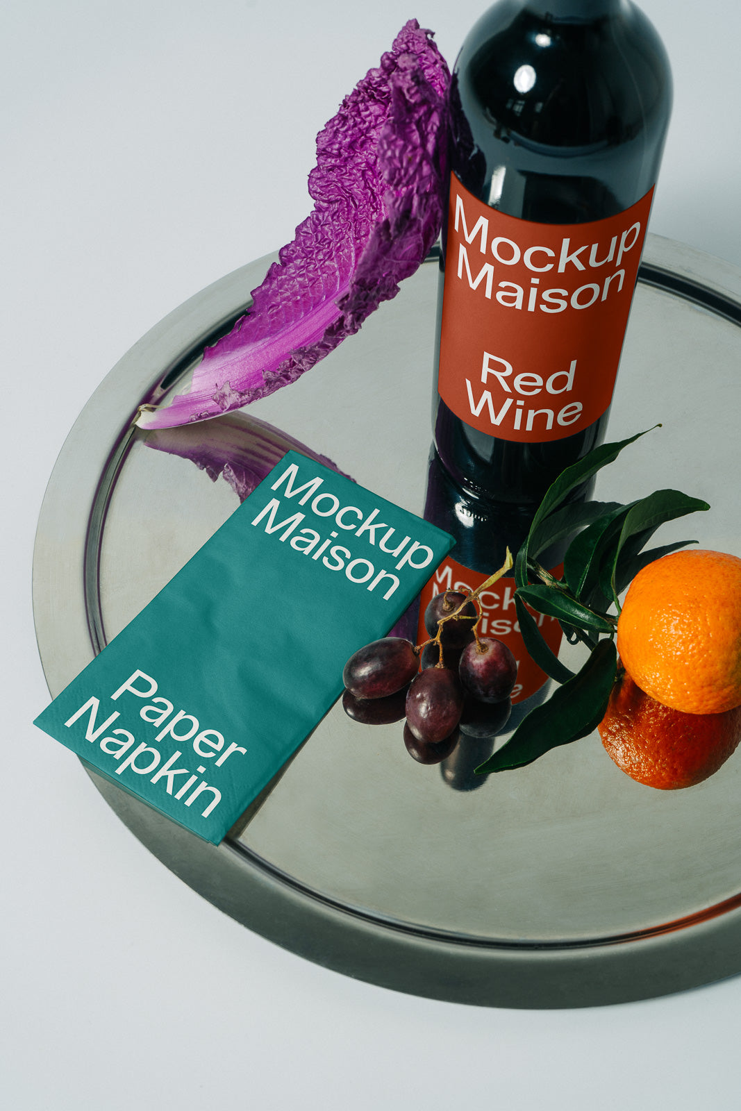 PSD mockup of wine bottle with red label beside teal paper napkin, placed on a metal tray with grapes, an orange, purple cabbage, and green leaves.