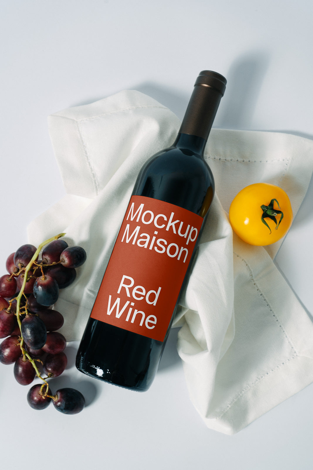 PSD mockup of a wine bottle labeled "Mockup Maison Red Wine," placed on a white cloth with red grapes and a yellow tomato nearby.