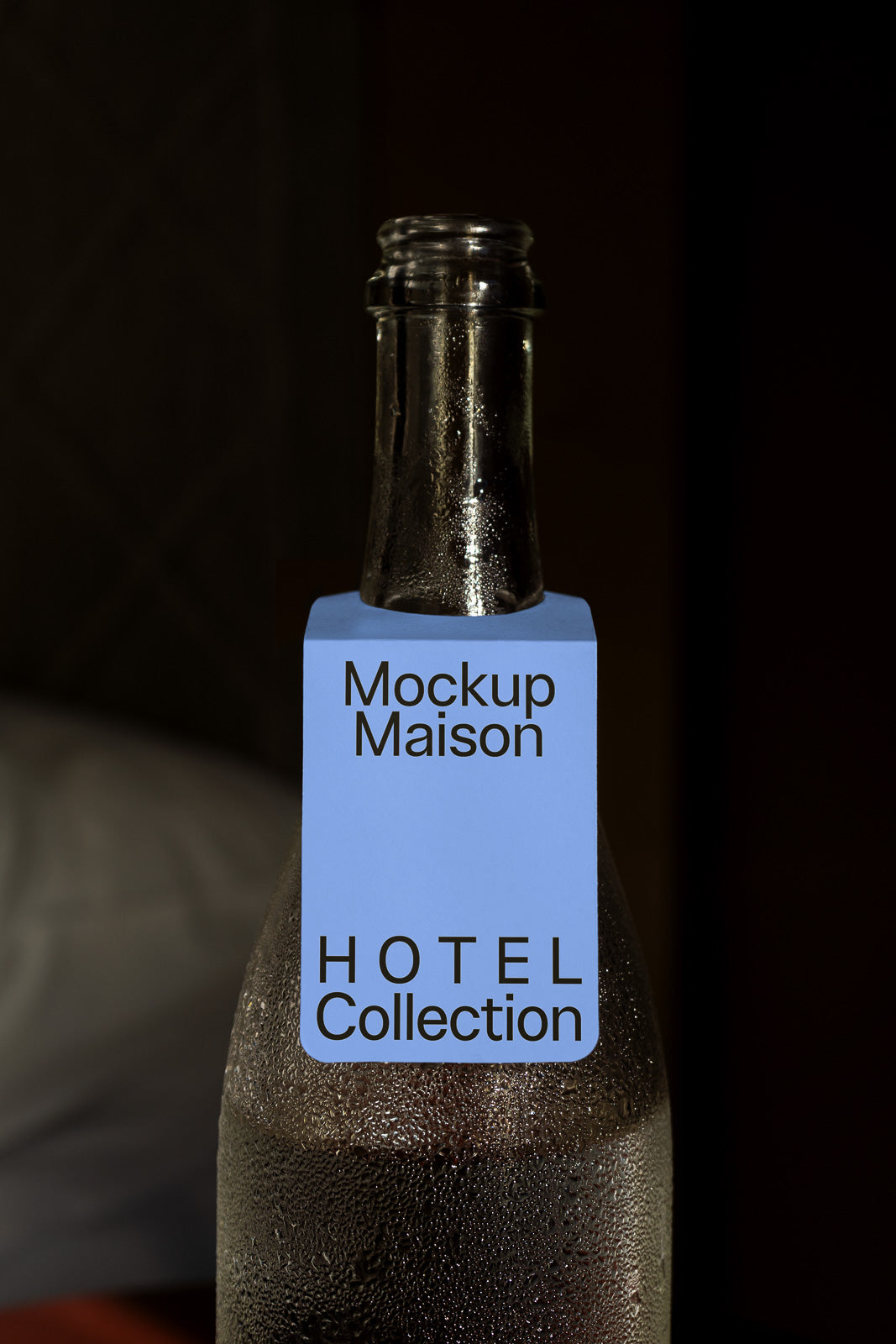 PSD mockup of a frosted glass bottle with a blue label tagged "Mockup Maison" and "HOTEL Collection," placed against a dark background.