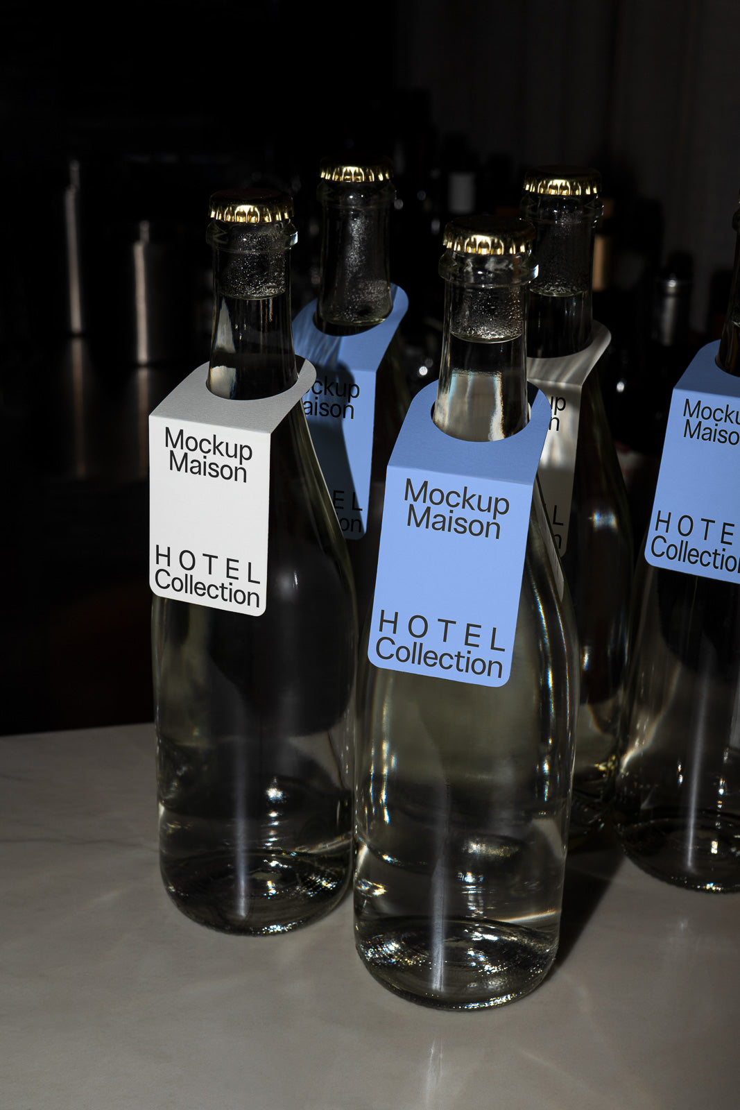 PSD mockup of glass bottles with gold caps and tags labeled “Mockup Maison HOTEL Collection” in a dimly lit setting.