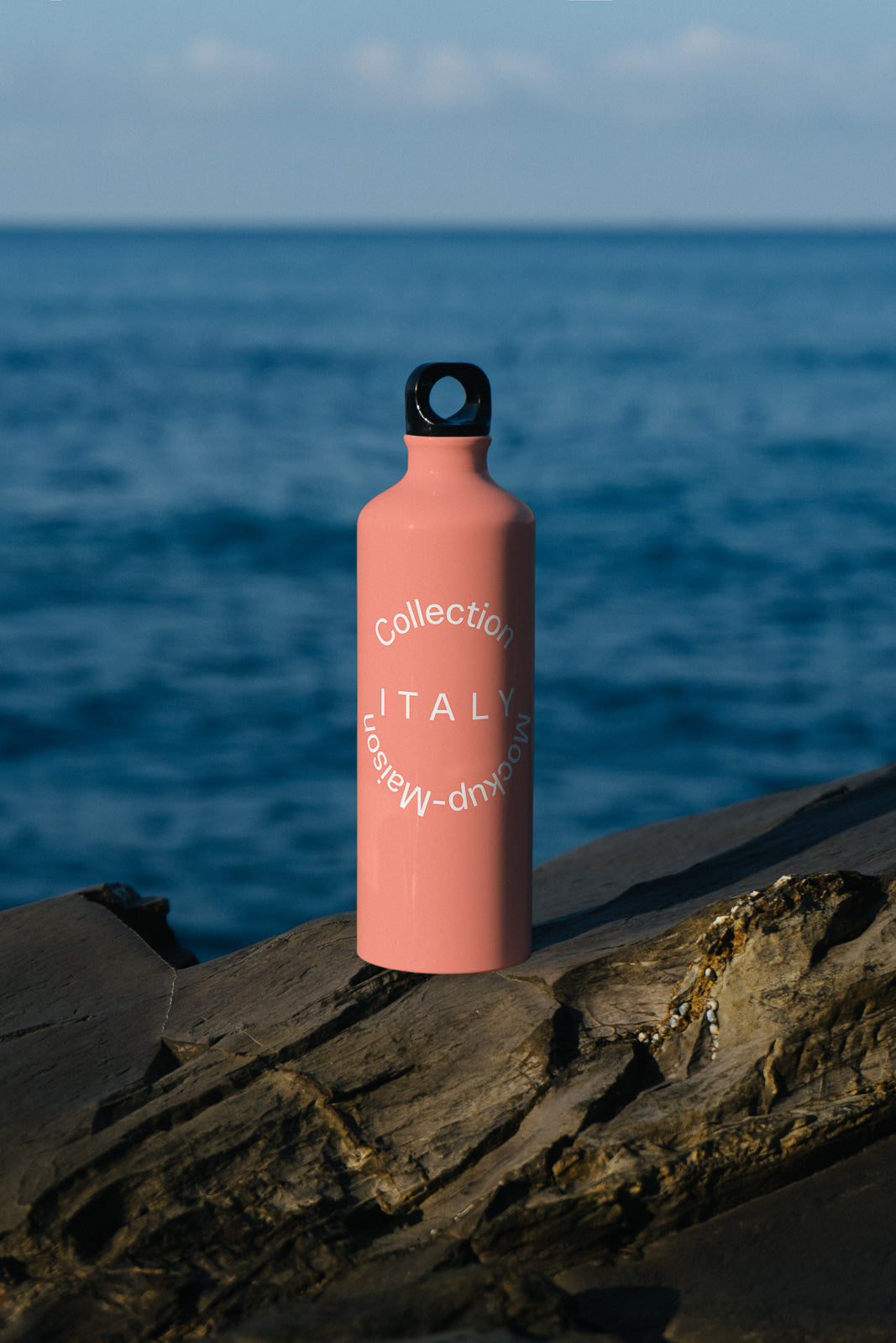 PSD mockup of a pink water bottle labeled "Collection Italy" by Mockup Maison, standing on a rock with a blurred ocean background.