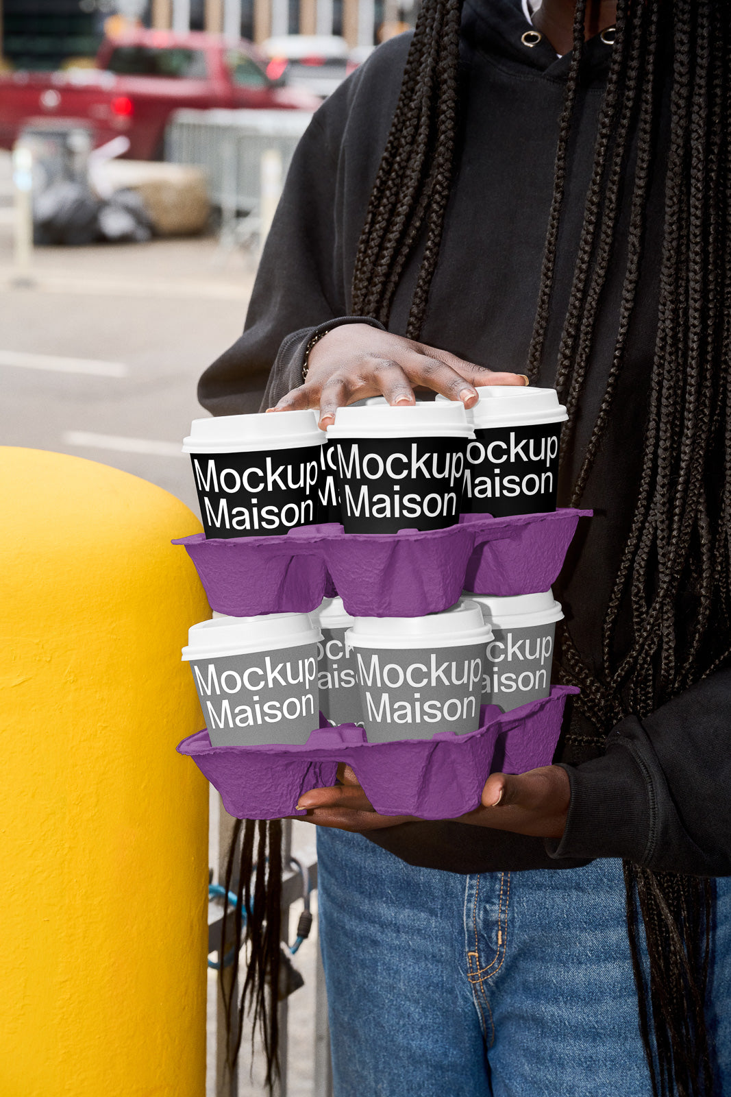 PSD mockup of a person holding two sets of takeaway coffee cups labeled "Mockup Maison" in a purple carrier, set against an urban background.