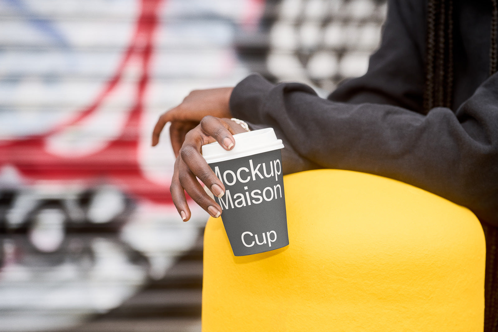 PSD mockup of a hand holding a coffee cup labeled "Mockup Maison Cup" against a blurred urban background, highlighting the product design.