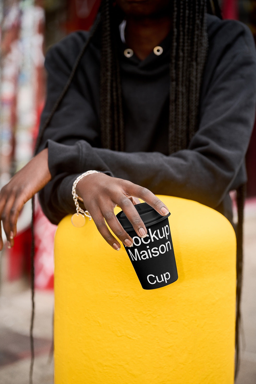 PSD mockup of a black coffee cup held by a person in a dark hoodie, resting on a bright yellow surface, with blurred urban background.