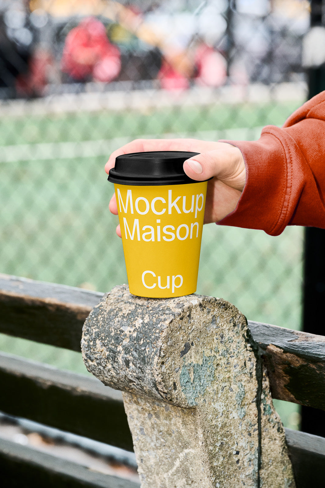 PSD mockup of a yellow coffee cup with black lid held by a person in a red sleeve, positioned on a weathered stone surface with a blurred fence background.