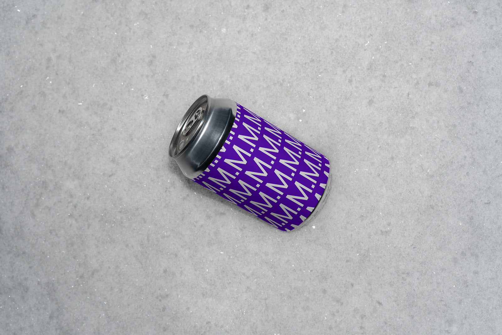 PSD mockup of a beverage can with a purple pattern featuring repeated "M" letters, placed on a textured, light gray surface.
