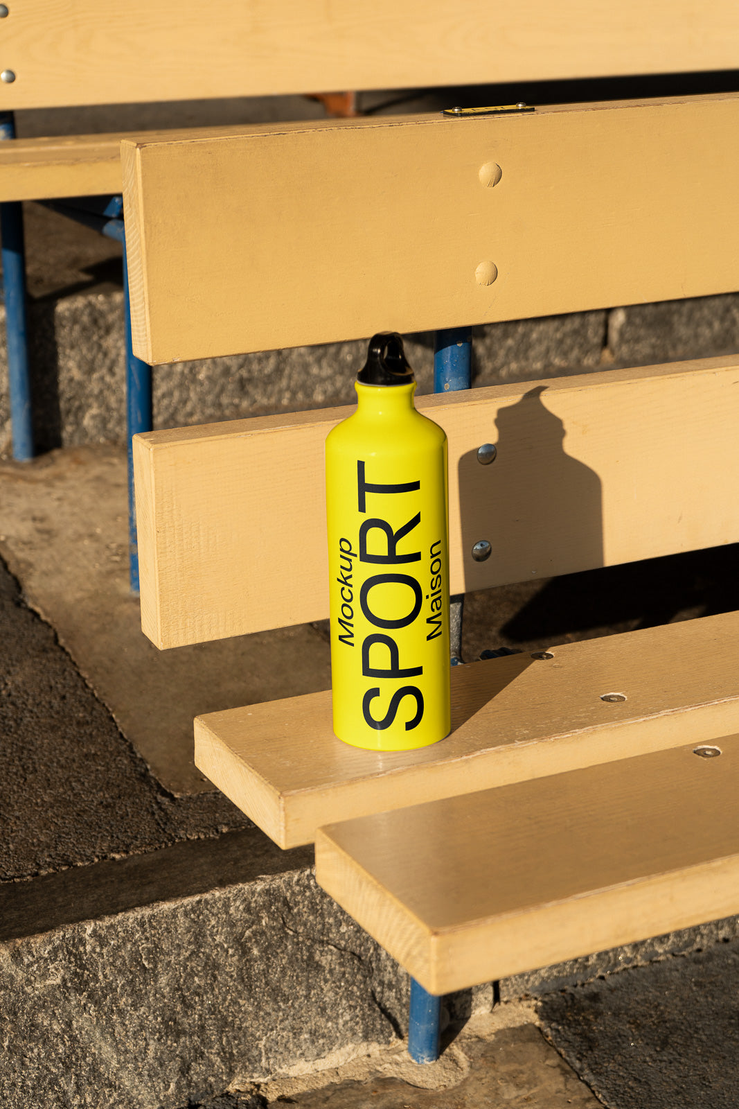 PSD mockup of a yellow sports bottle labeled "Maison" resting on wooden benches outdoors, casting a shadow in warm sunlight.