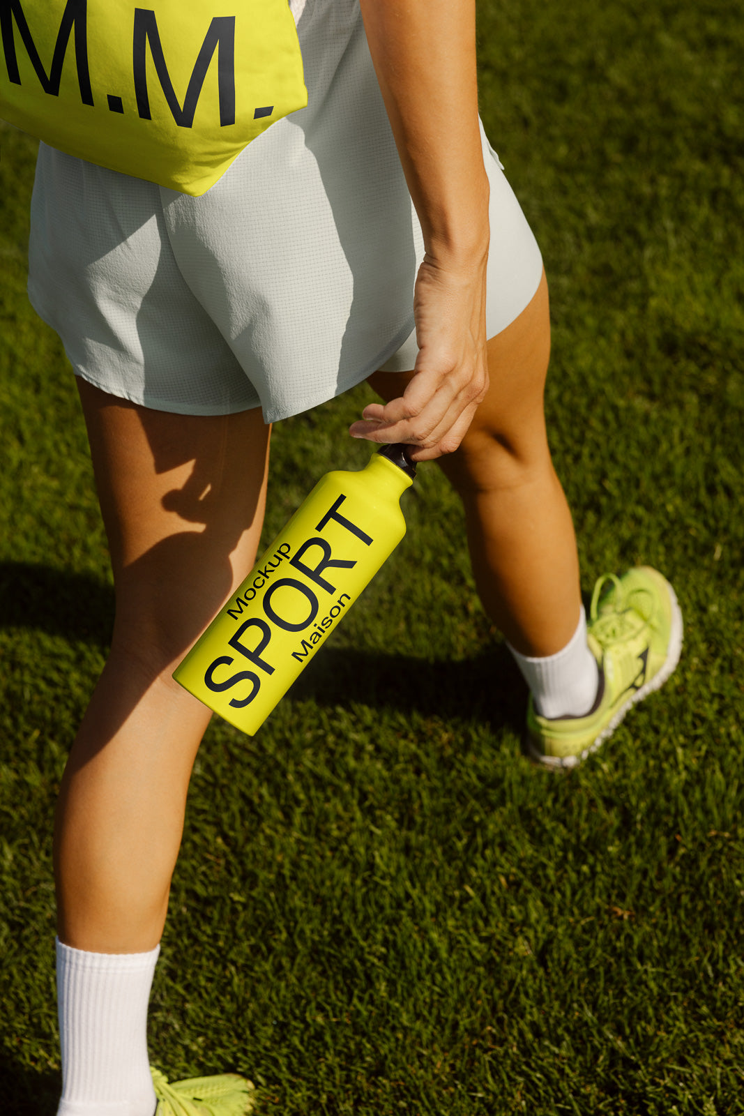 PSD mockup of a person holding a neon yellow sports bottle labeled "Mockup Sport Maison," standing on a grassy field, dressed in athletic wear.