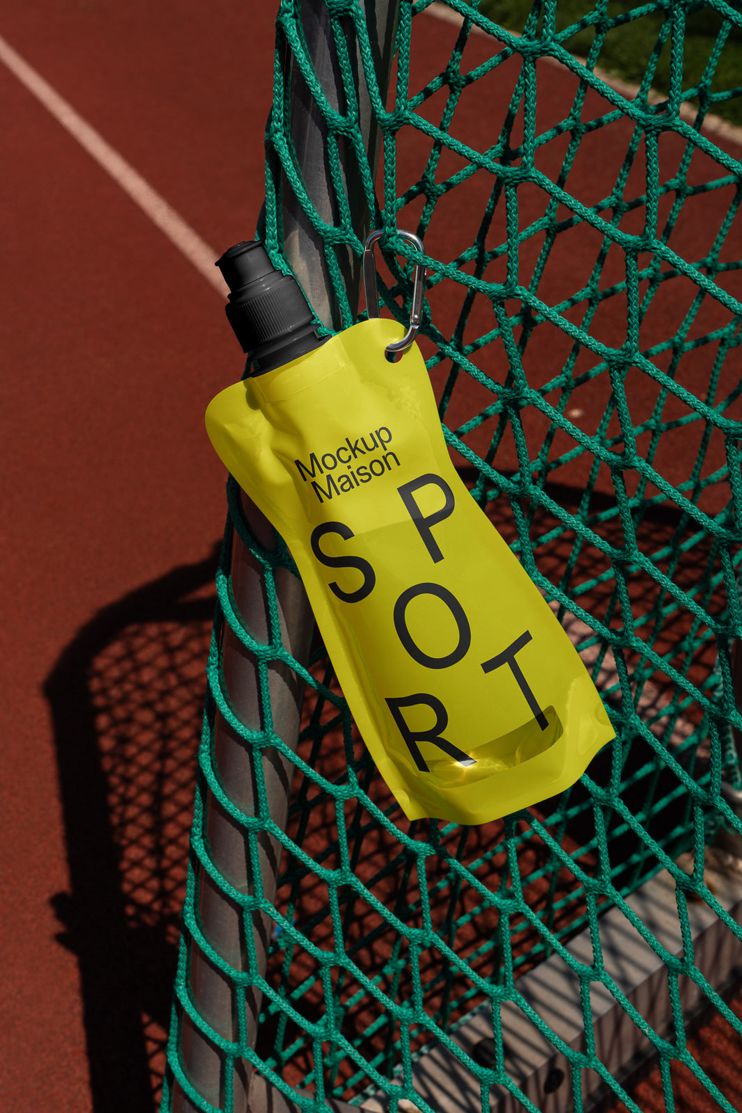 PSD mockup of a yellow sports pouch with a black cap hanging on a green net near a red running track, labeled "Mockup Maison SPORT."