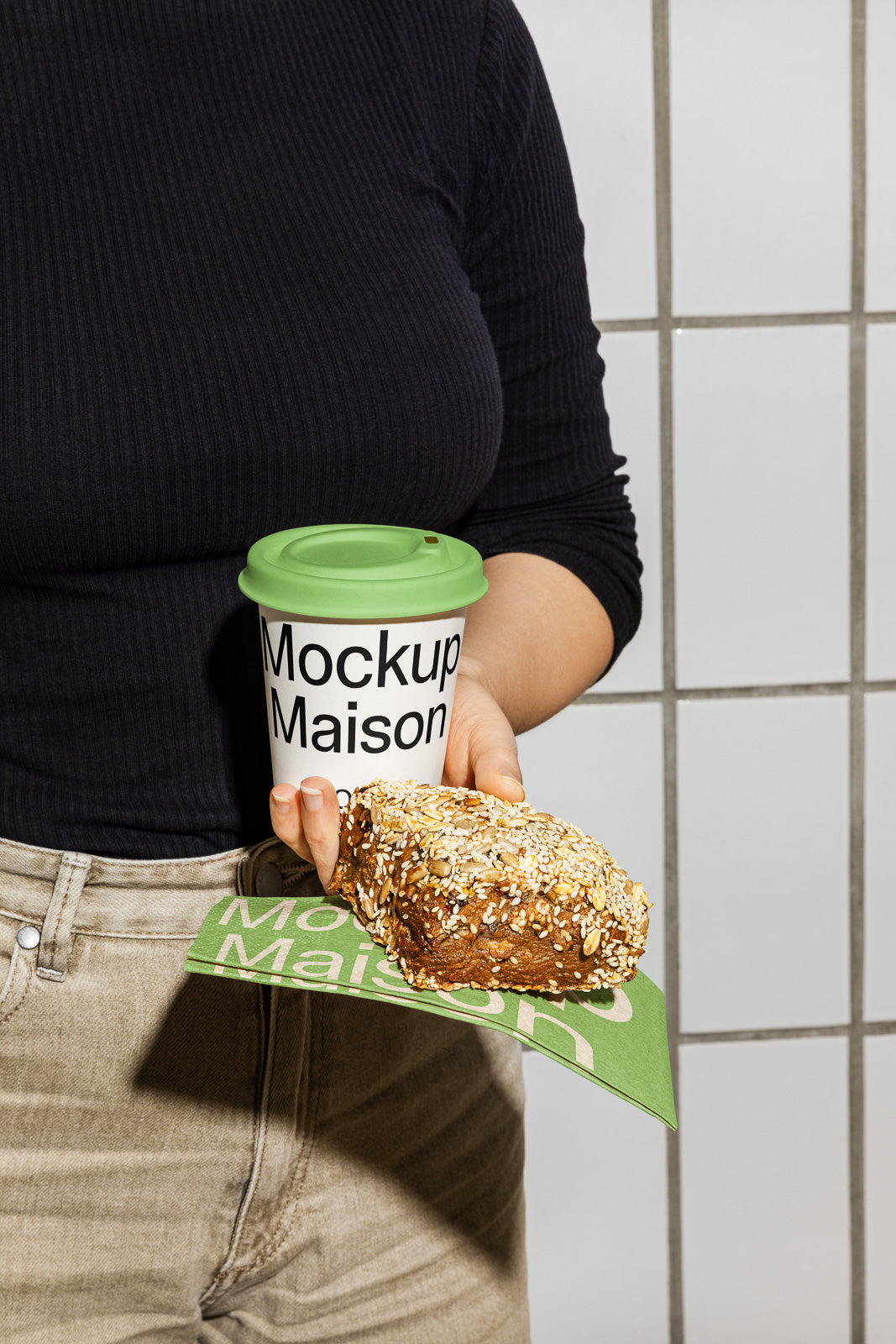 PSD mockup of a person holding a coffee cup with a green lid labeled "Mockup Maison" and a seeded bread roll on a green napkin.
