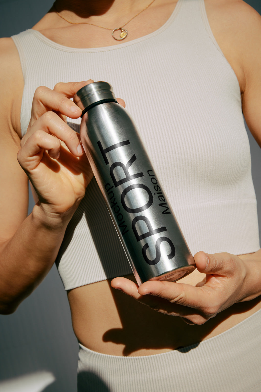 PSD mockup of a person holding a metallic water bottle labeled "Sport Maison" against a light background, showcasing the product design in a casual setting.