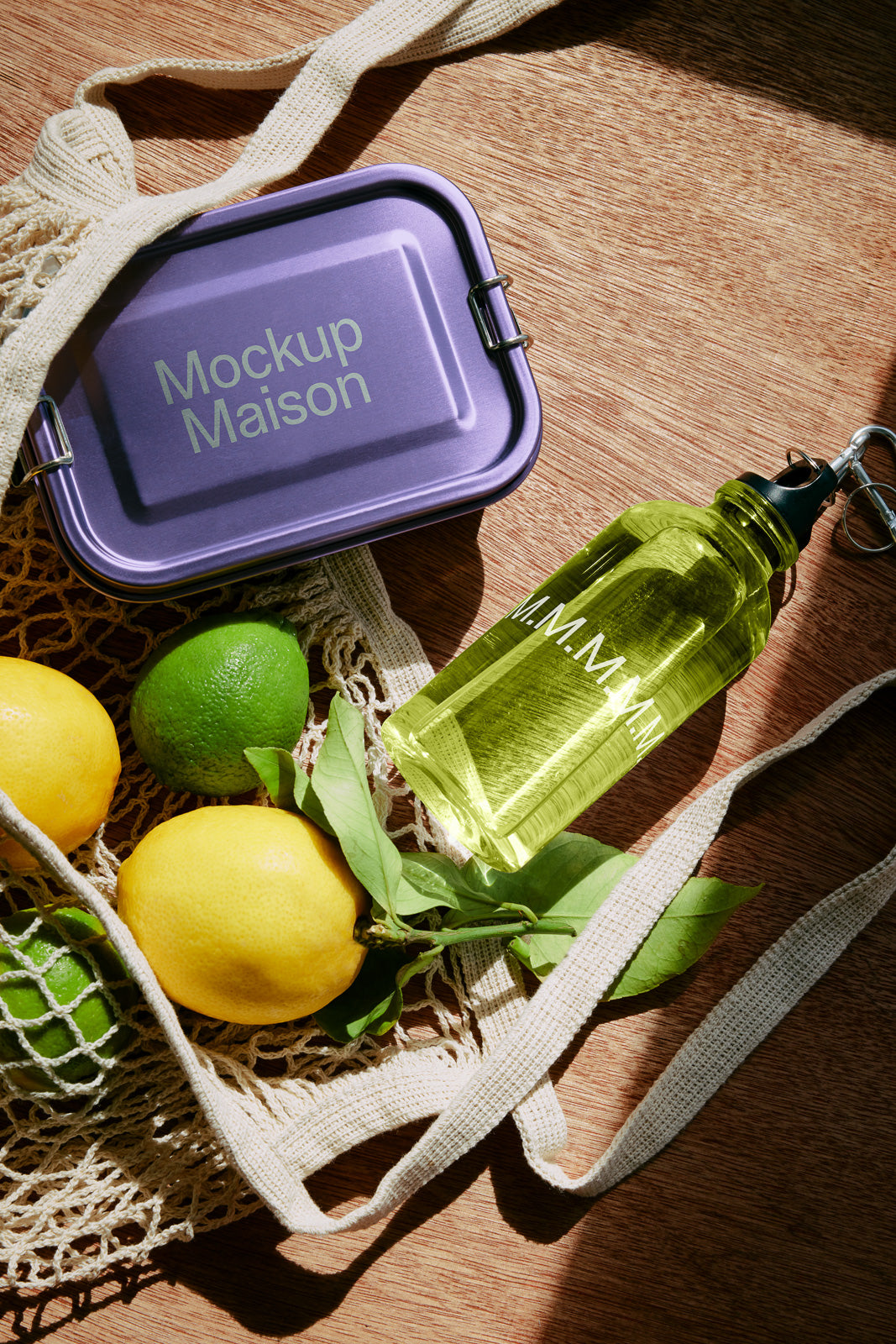PSD mockup of a purple lunchbox with "Mockup Maison" text, laying on a wooden surface next to a green water bottle and a net bag holding lemons and limes.