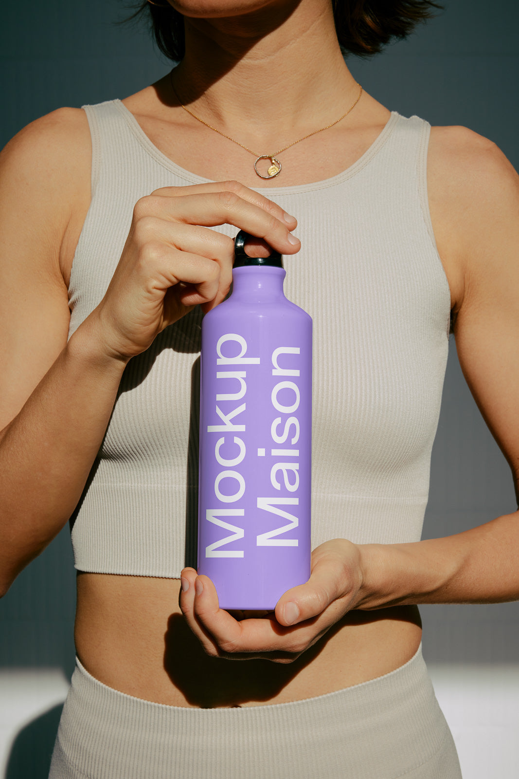 PSD mockup of a person holding a purple water bottle with the words "Mockup Maison" printed vertically. The person wears a light beige tank top.