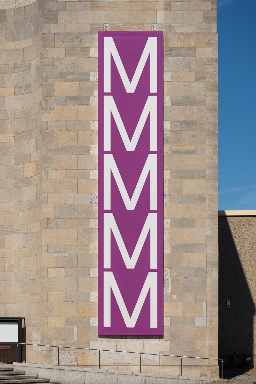 PSD mockup of a large vertical banner with repeated white letter M on a purple background, hung on a brick exterior wall.
