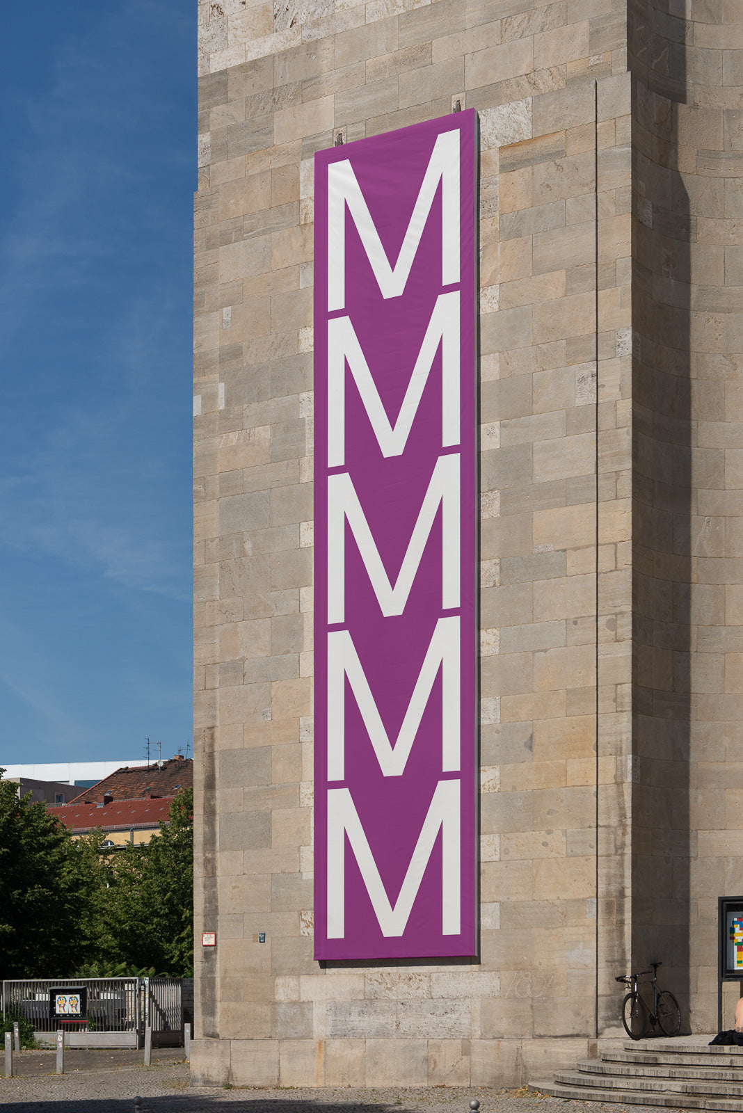 PSD mockup of large purple banner with repeated white letter "M" affixed to a stone building facade.
