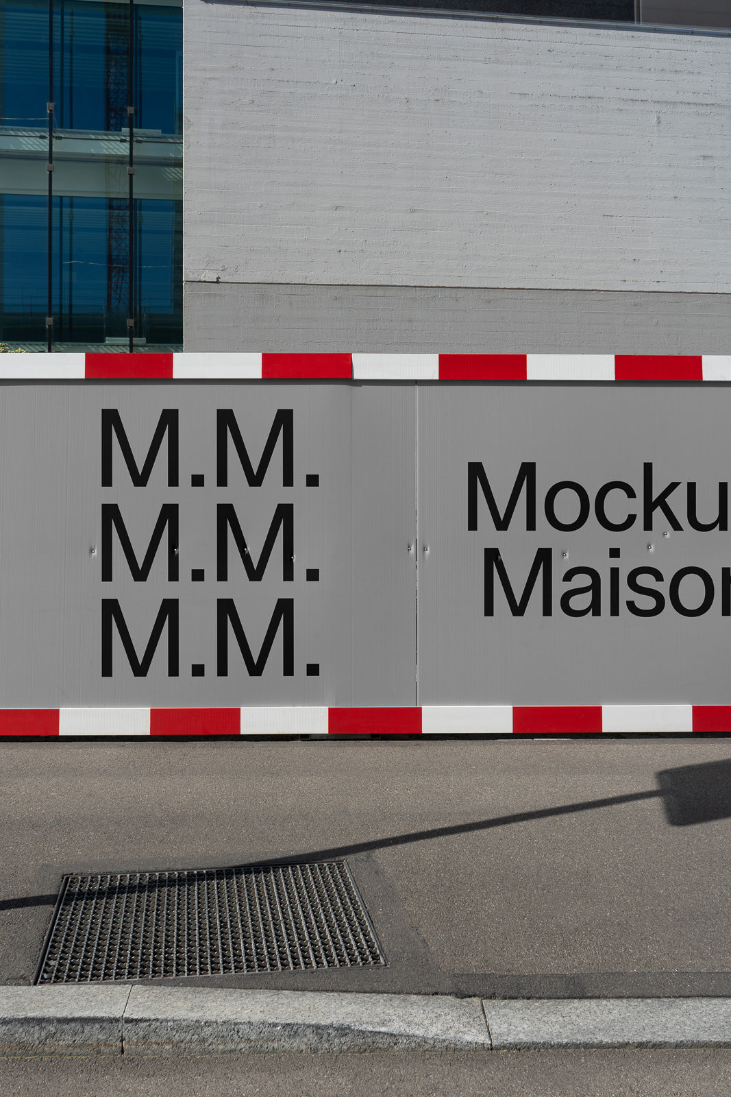 PSD mockup of an outdoor construction site barrier with bold black text "M.M." in multiple rows and "Mockup Maison" on a gray background.