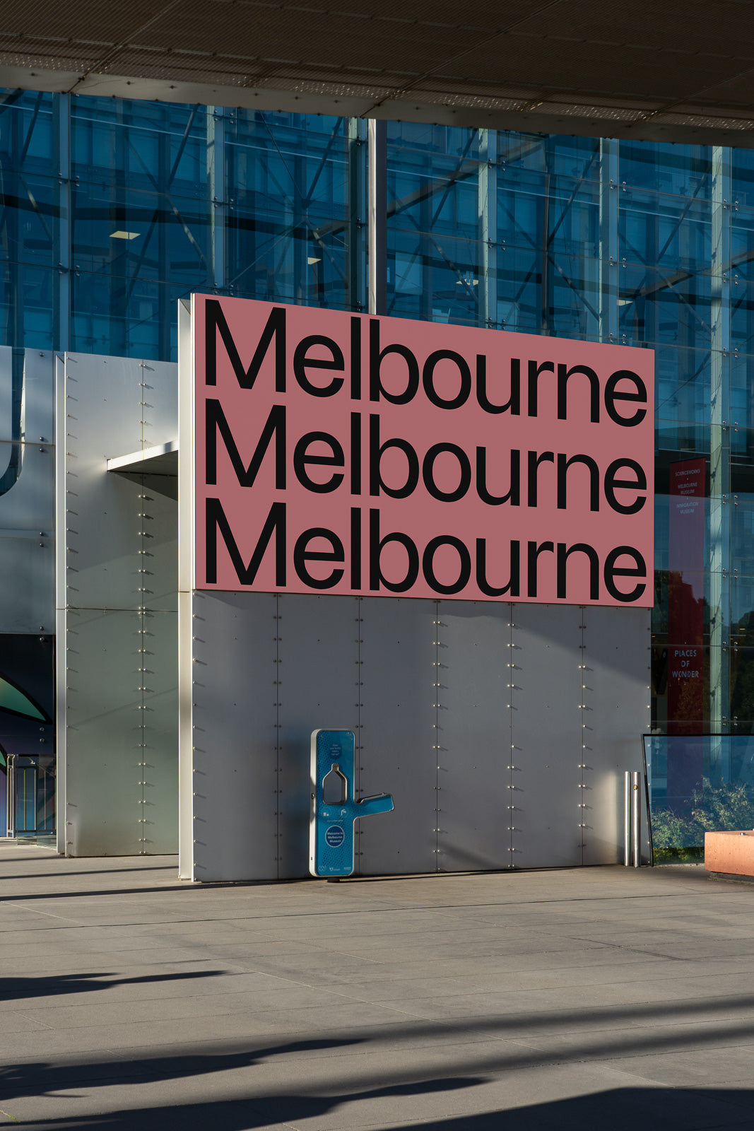 PSD mockup of a large outdoor signage display with the word "Melbourne" repeated three times in bold, black letters on a pink background.