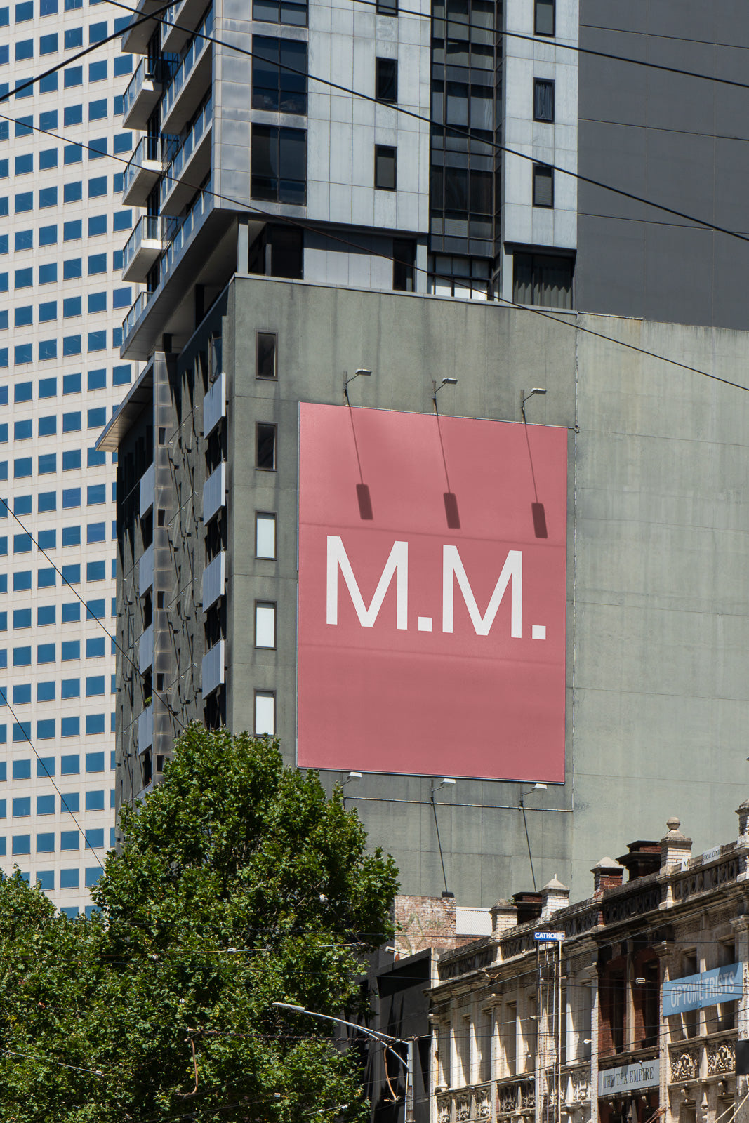 PSD mockup of a large pink billboard with "M.M." text on the side of a tall urban building, surrounded by modern architecture and trees.