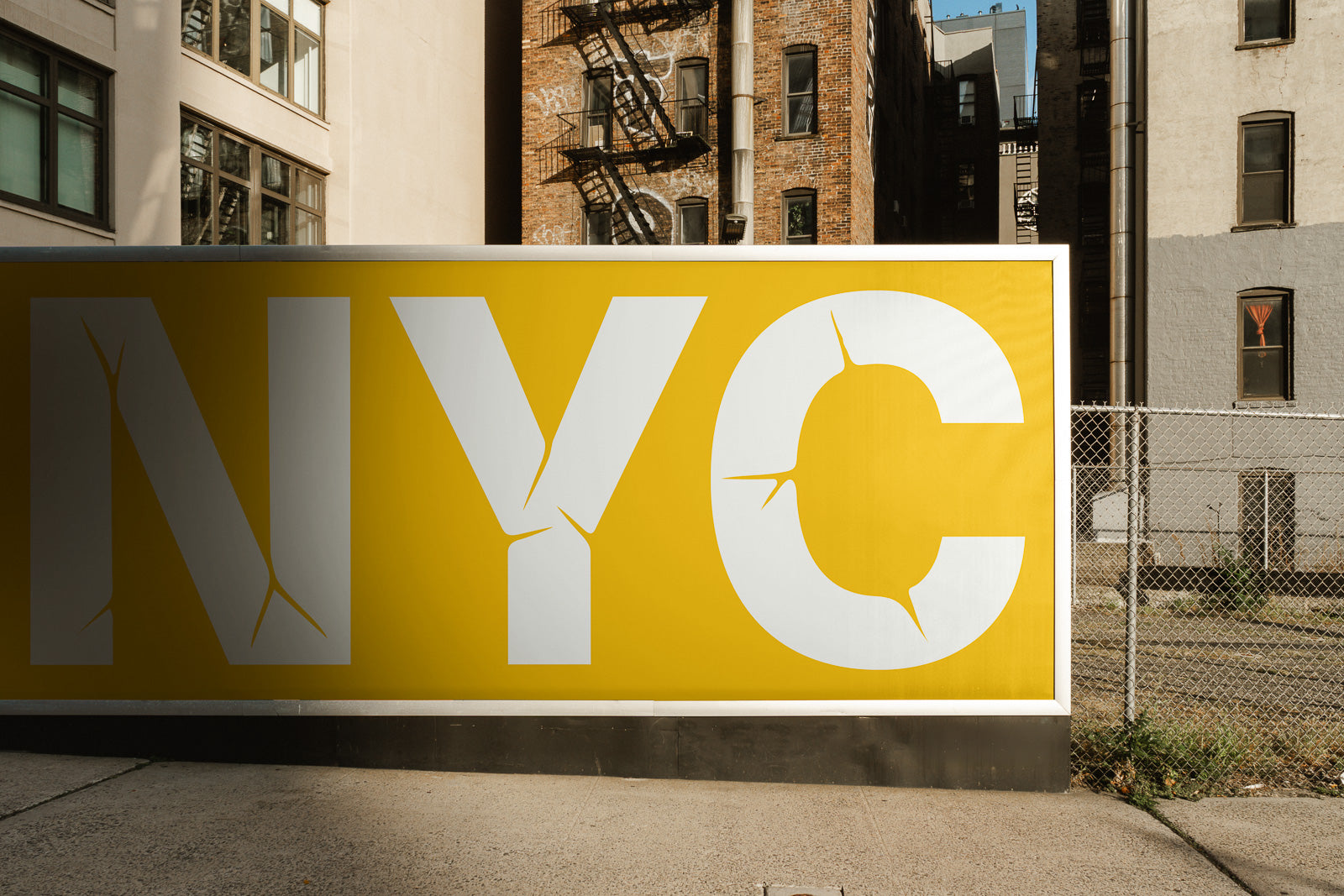 PSD mockup of a large yellow billboard with cracked-style "NYC" letters, set against an urban background with buildings and a chain-link fence.