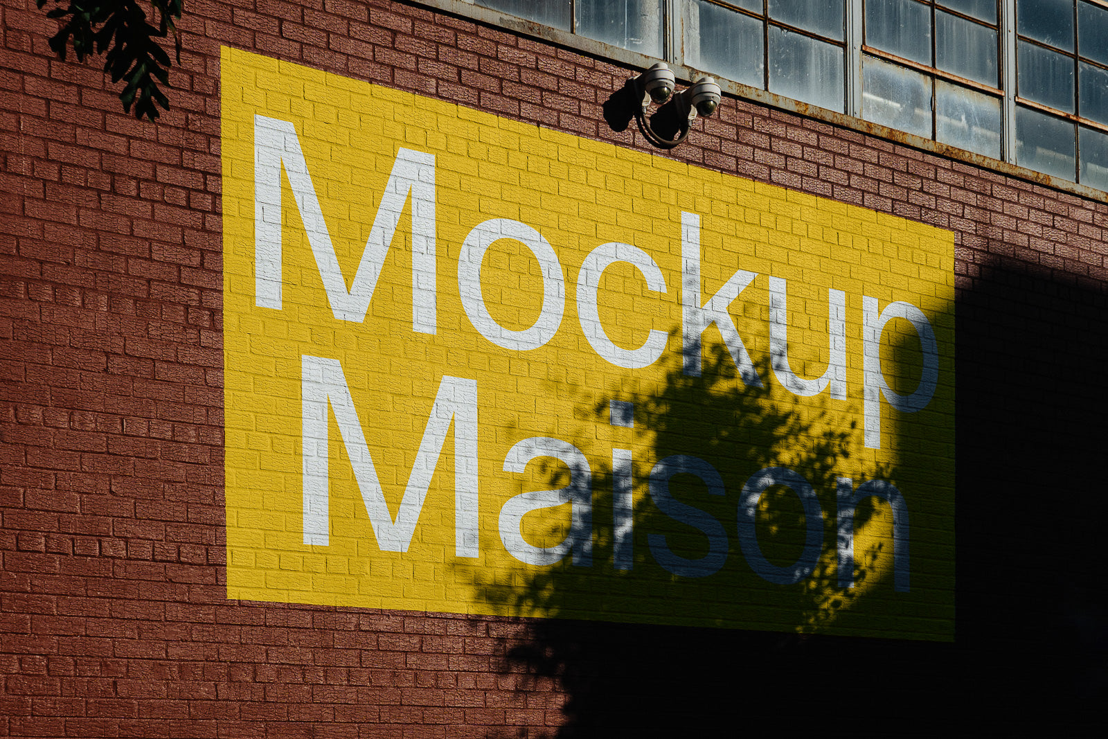 PSD mockup of a yellow advertisement on a brick wall with the text "Mockup Maison," partially shaded by a tree, featuring windows above.