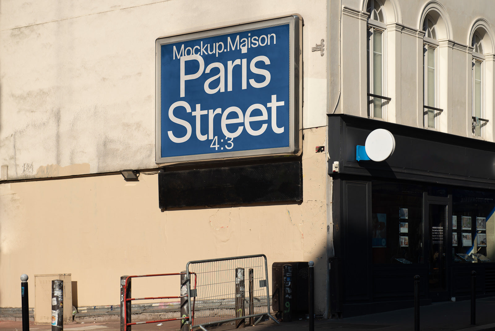 PSD mockup of a street with a blue rectangular sign displaying "Mockup.Maison Paris Street 4:3" on a light-colored wall adjacent to a storefront.