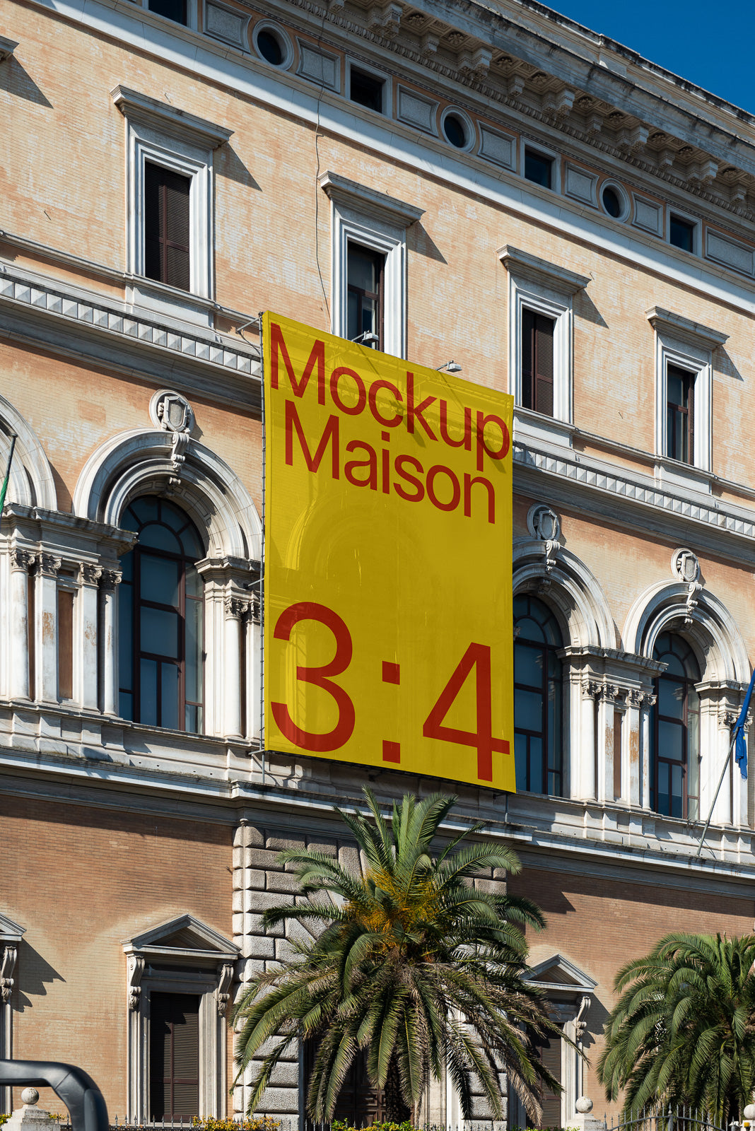 PSD mockup of a large yellow banner displaying "Mockup Maison 3:4" on a historic building facade with arched windows and palm trees below.
