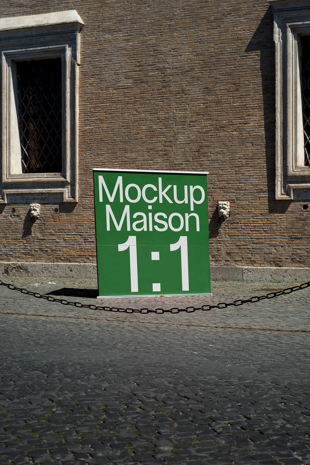 PSD mockup of a large green sign reading 'Mockup Maison 1:1' placed against a historic brick wall with barred windows, surrounded by cobblestones.