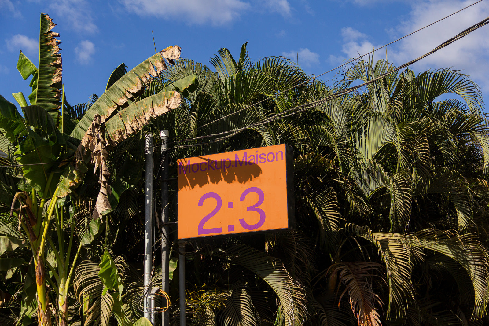PSD mockup of a rectangular orange sign with purple text "Mockup Maison 2:3" against a backdrop of lush palm trees and a clear blue sky.