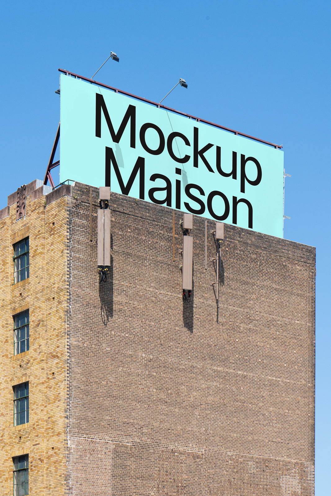 PSD mockup of a large outdoor billboard mounted on a brick building, displaying text "Mockup Maison" against a clear blue sky.