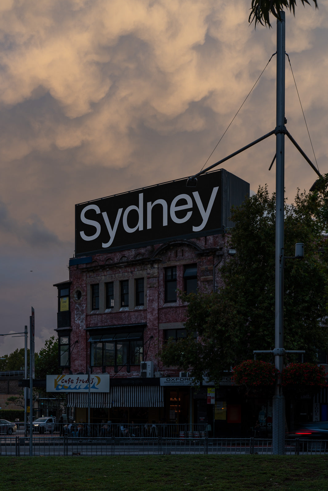 PSD mockup of a vintage brick building with a large "Sydney" sign, set against a dramatic, cloudy sky with subtle lighting from streetlamps below.