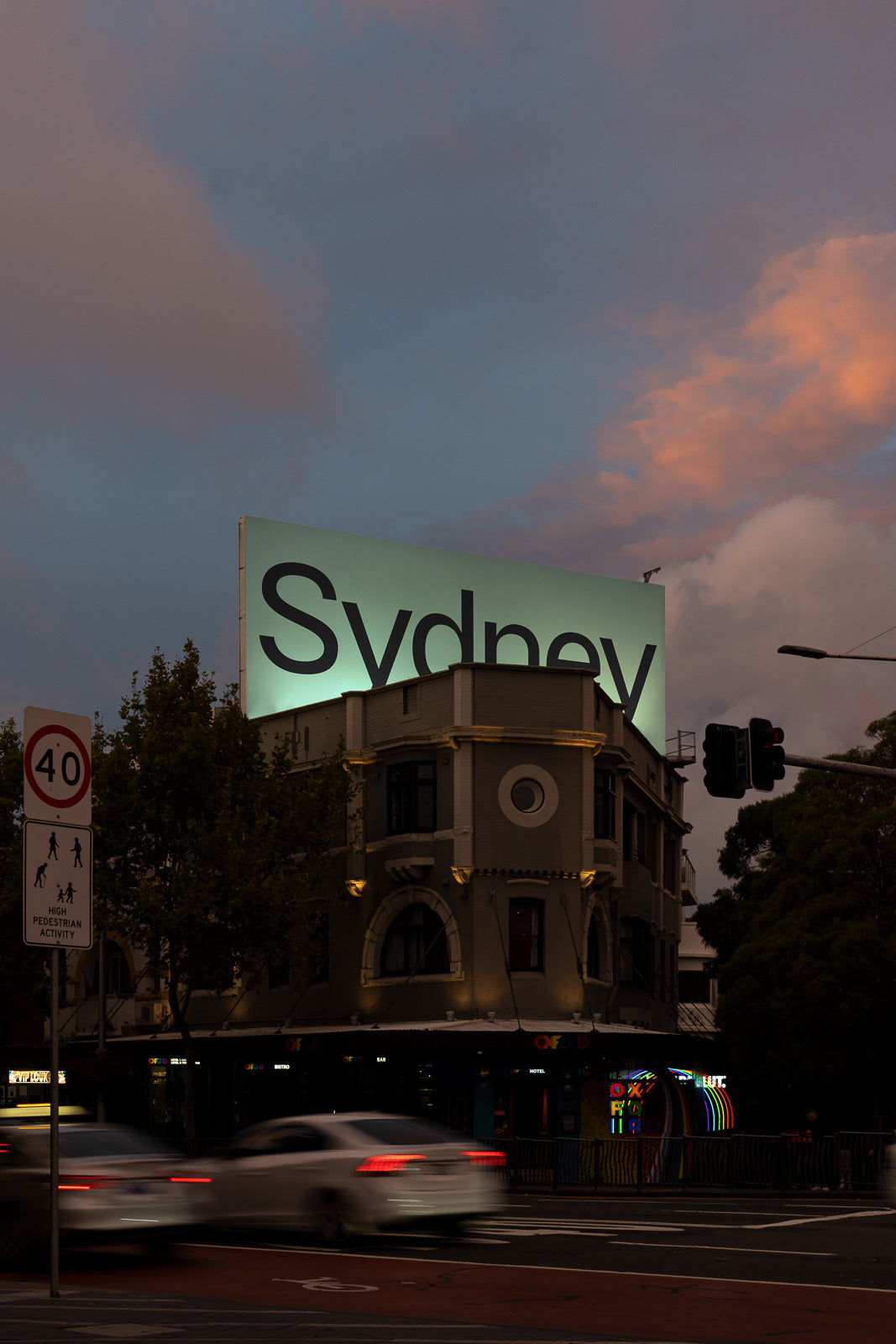 PSD mockup of a large illuminated billboard displaying the word "Sydney" atop a historic building, with blurred cars passing by on a city street at dusk.