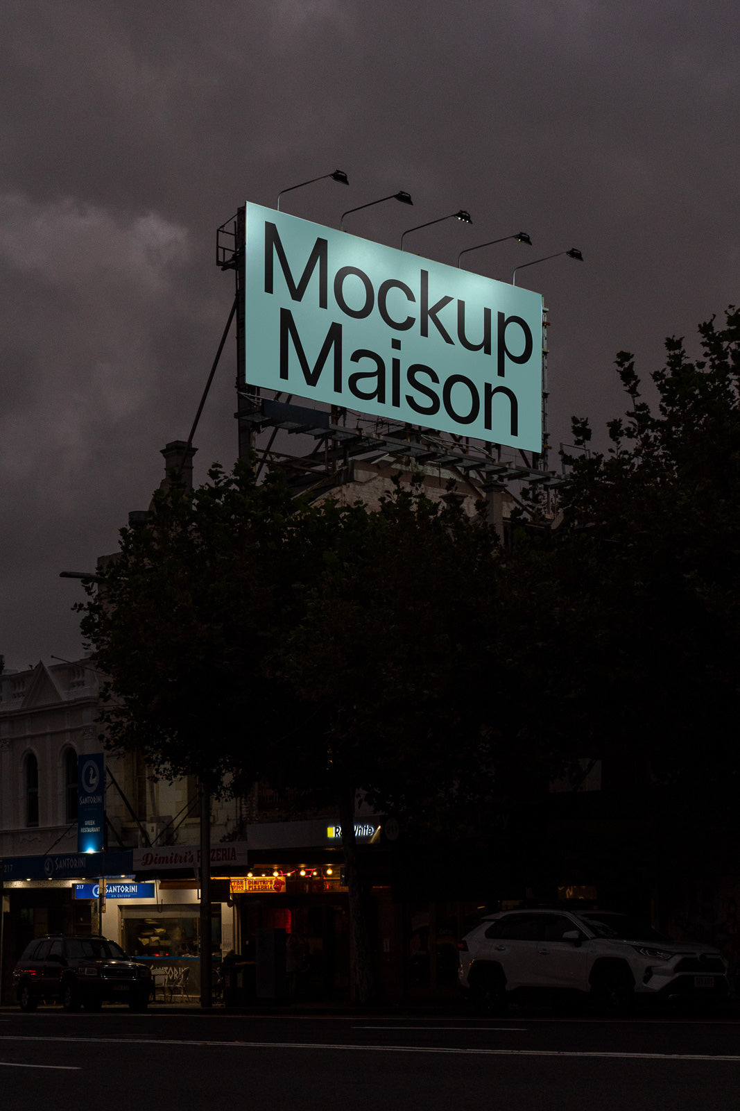 PSD mockup of a large illuminated billboard displaying "Mockup Maison" against a dark, cloudy evening sky, with city buildings and cars below.